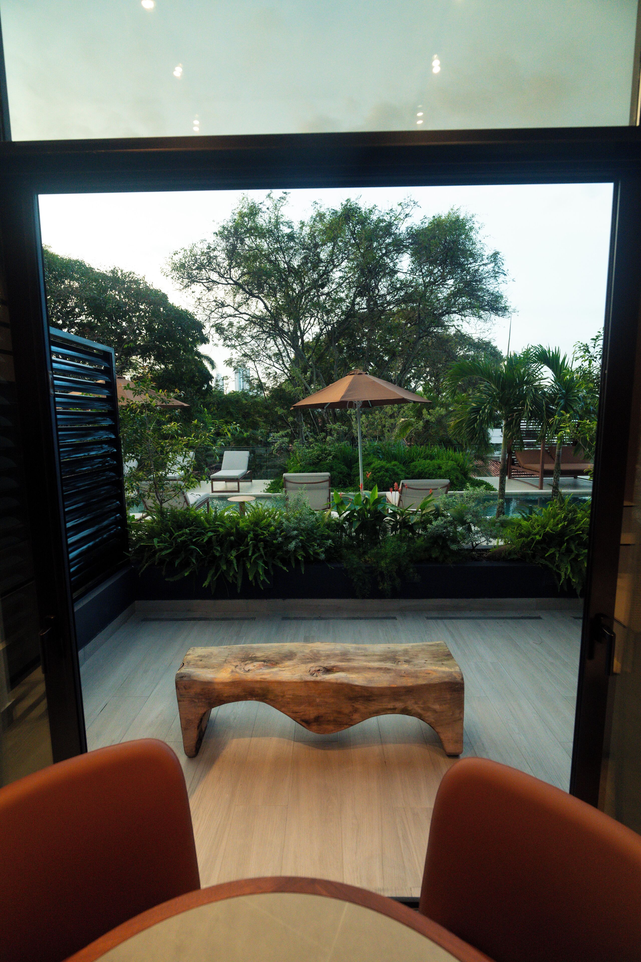 Un interior moderno con un banco de madera, dos sillas y una pared de vidrio que ofrece vistas de un jardín exterior con piscina y sombrillas.