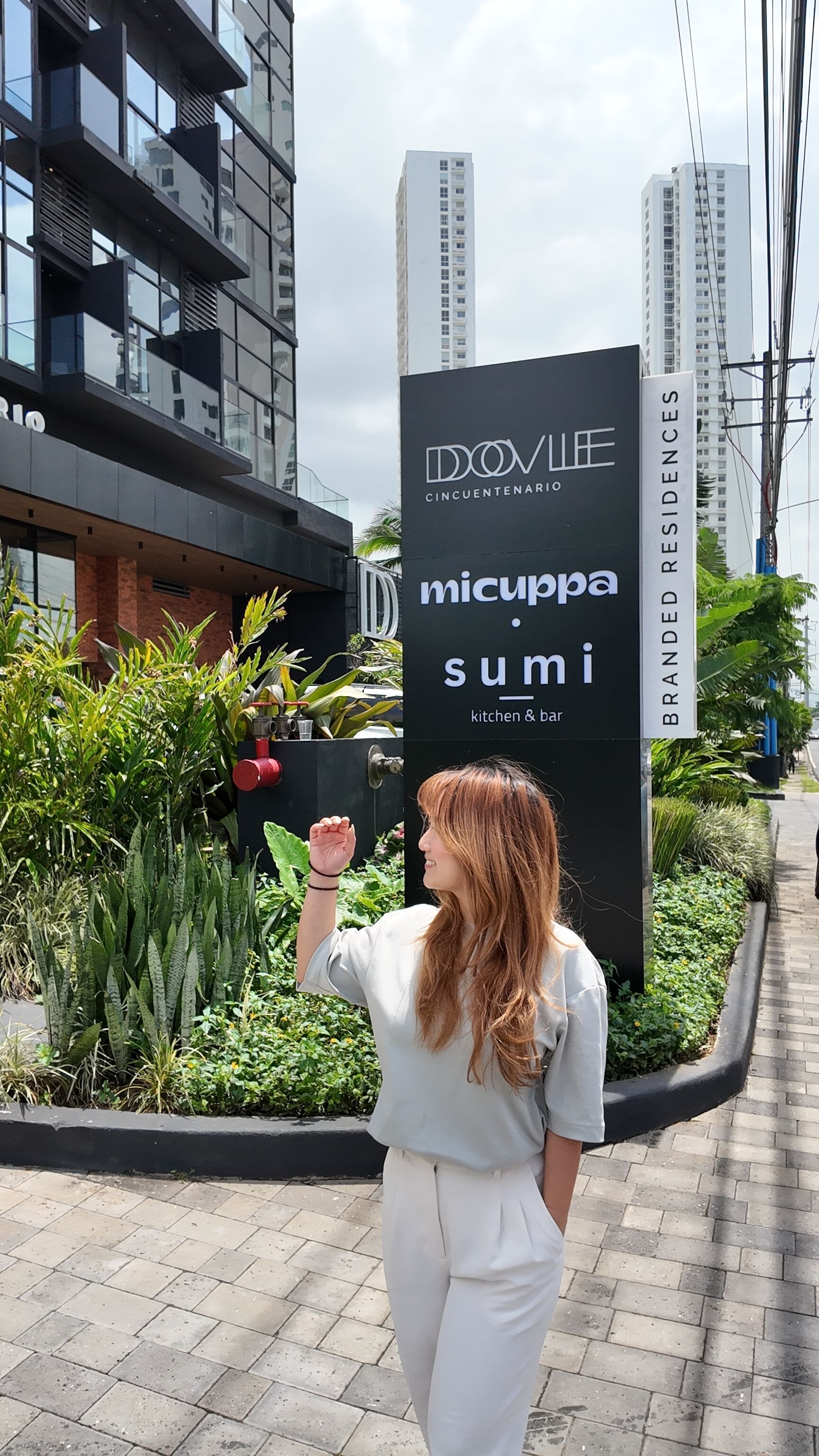 Una persona de pie frente a un letrero de 'micuppa sumi', con un edificio moderno al fondo.