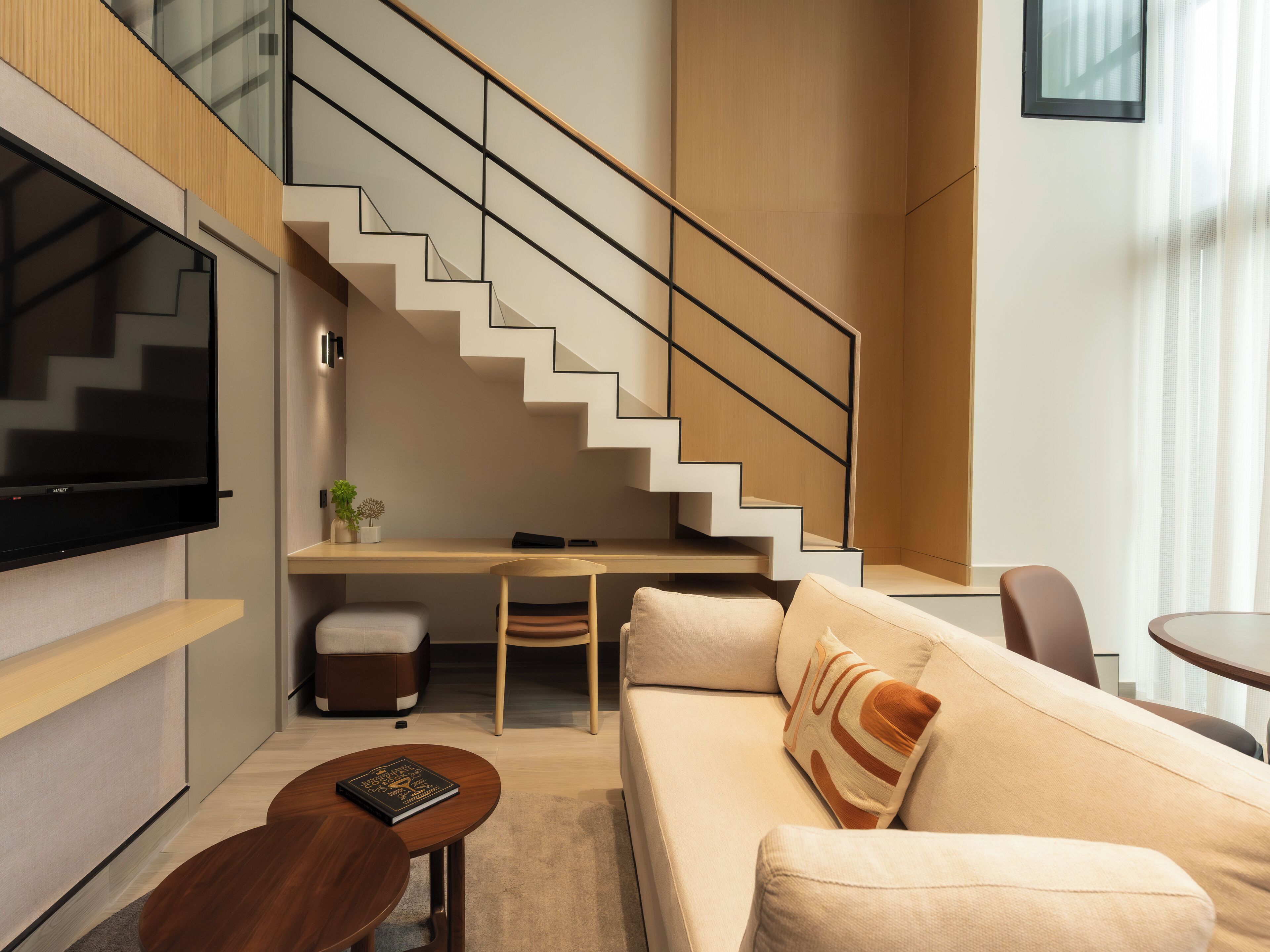 A modern living room with a staircase, a flat-screen TV, a desk, and a sofa.