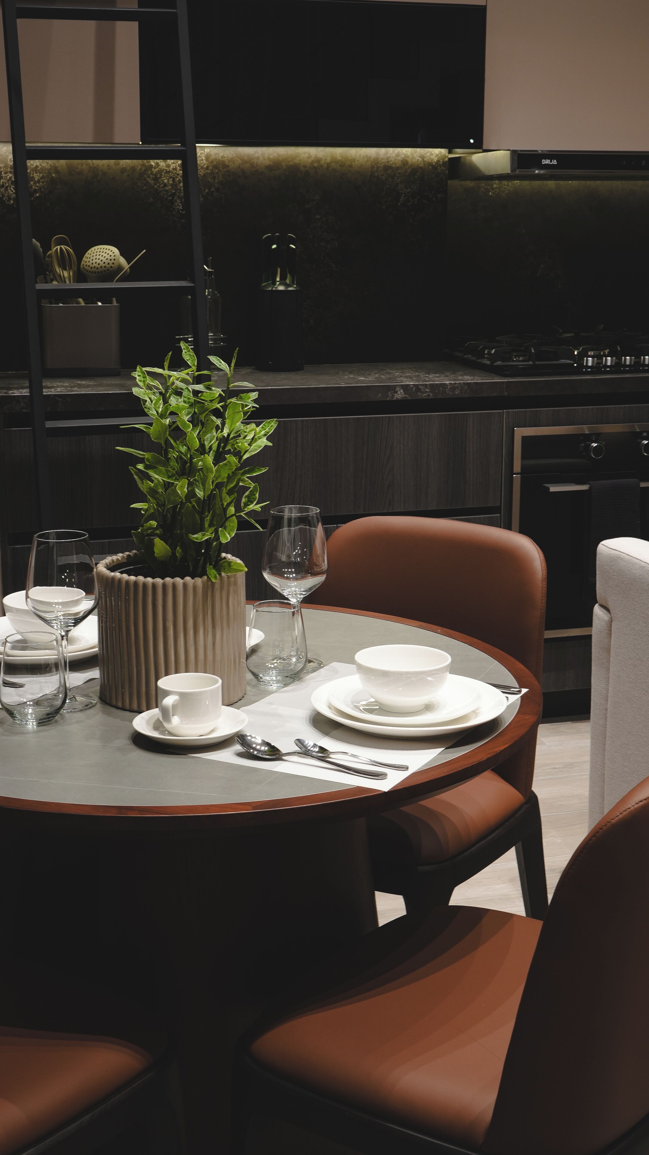 A modern dining area with a round table set for two, featuring white plates, glasses, and cutlery. A potted plant adds a touch of greenery.