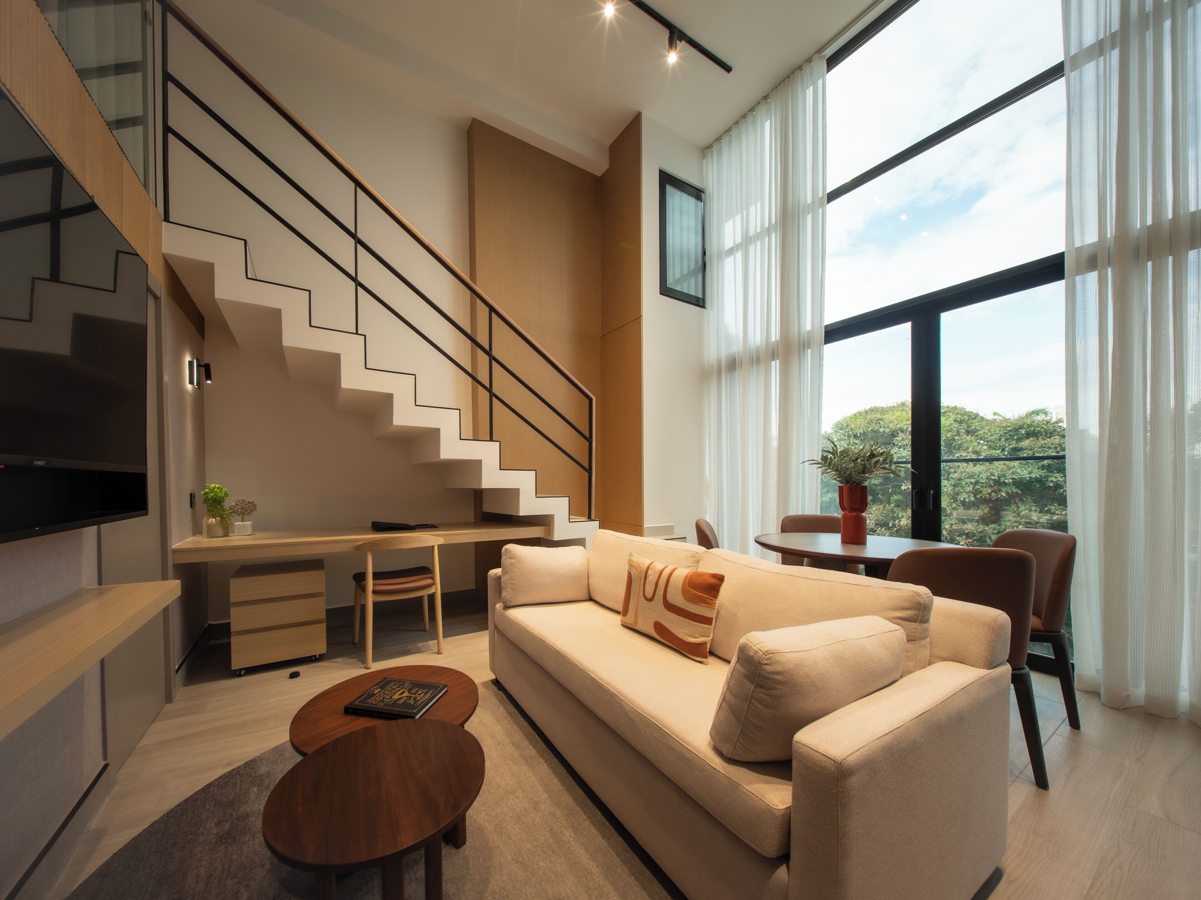 A modern living room with a staircase, a sofa, a coffee table, and a dining area with a view of greenery outside.