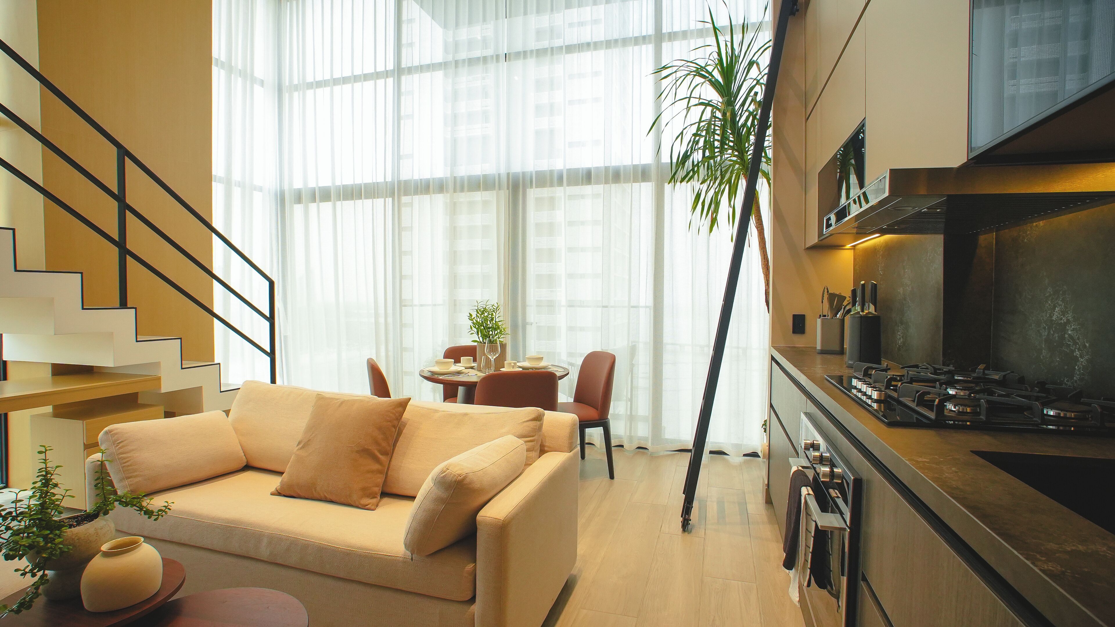 Un salon moderne avec un canapé, une table à manger et des chaises. Un escalier se trouve à gauche, et un coin cuisine avec une cuisinière et des placards se trouve à droite.