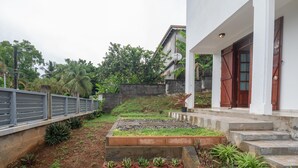 A garden area with raised beds and potted plants, a concrete pathway, and a white building with a red door.