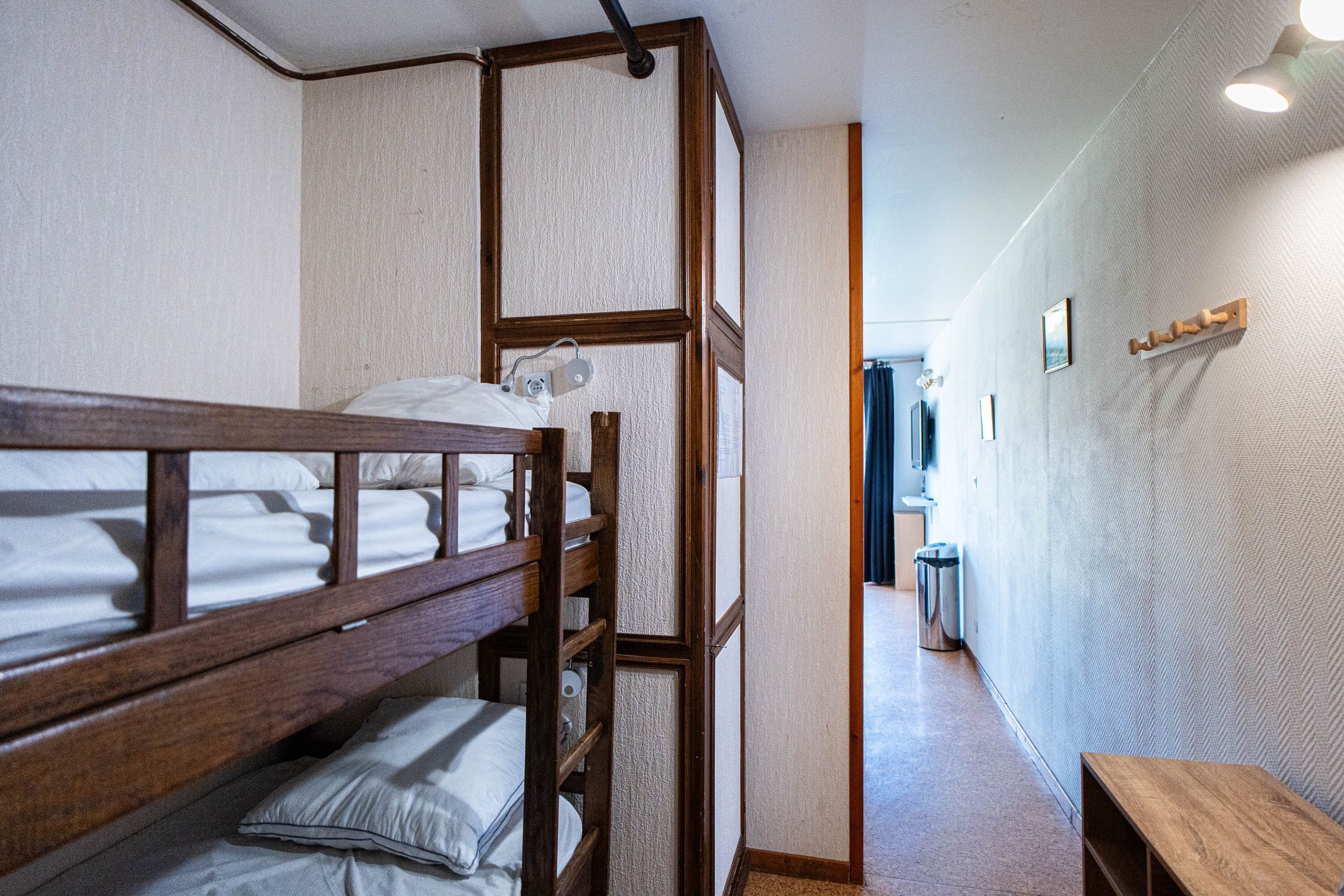 Quarto com beliche, cabeceira de madeira, um pequeno banco de madeira e um cesto de lixo no corredor.
