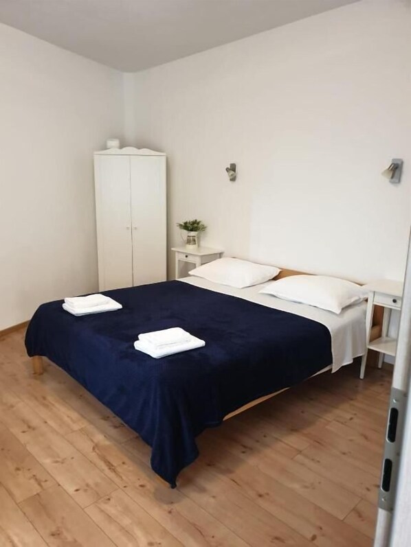 Un lit bien fait, recouvert d’une couvre-lit bleu foncé et agrémenté d’oreillers blancs, une armoire blanche et une petite table de chevet sur laquelle est posée une plante.