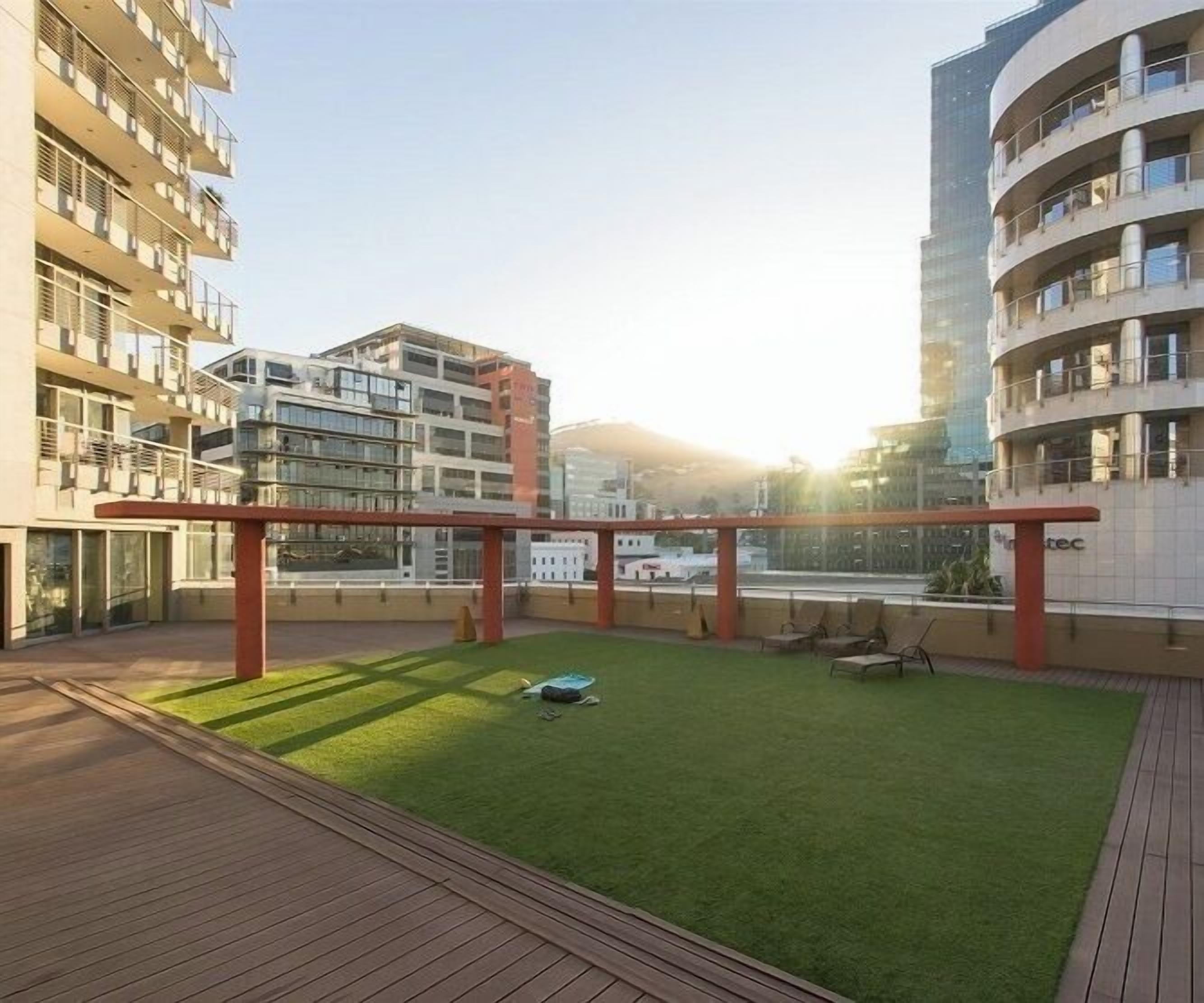 A rooftop garden with a wooden deck, a small lawn, and a pergola structure.