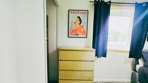 A room with a wooden dresser, a vintage poster on the wall, a window with blinds, and a curtain. - Comfy 3BR Flat on Quiet Street in Wollaton with FREE Parking (Nottingham)
