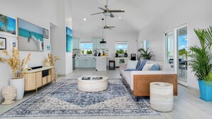 Een moderne woonkamer met een bank, salontafel en een groot vloerkleed. Achtergrond toont een keuken met lichtblauwe kasten en een eethoek met tafel en stoelen.