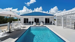 Een zwembad met witte ligbedden en parasols, een overdekt terras en een gebouw met een blauw dak.