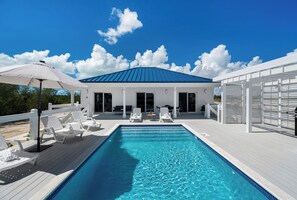 Una piscina con poltrona a sdraio bianche e ombrelloni, una zona patio coperta e un edificio con tetto blu.