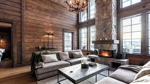Acogedor interior de cabaña de madera con chimenea de piedra, sofá modular y un ventanal con vistas al exterior.