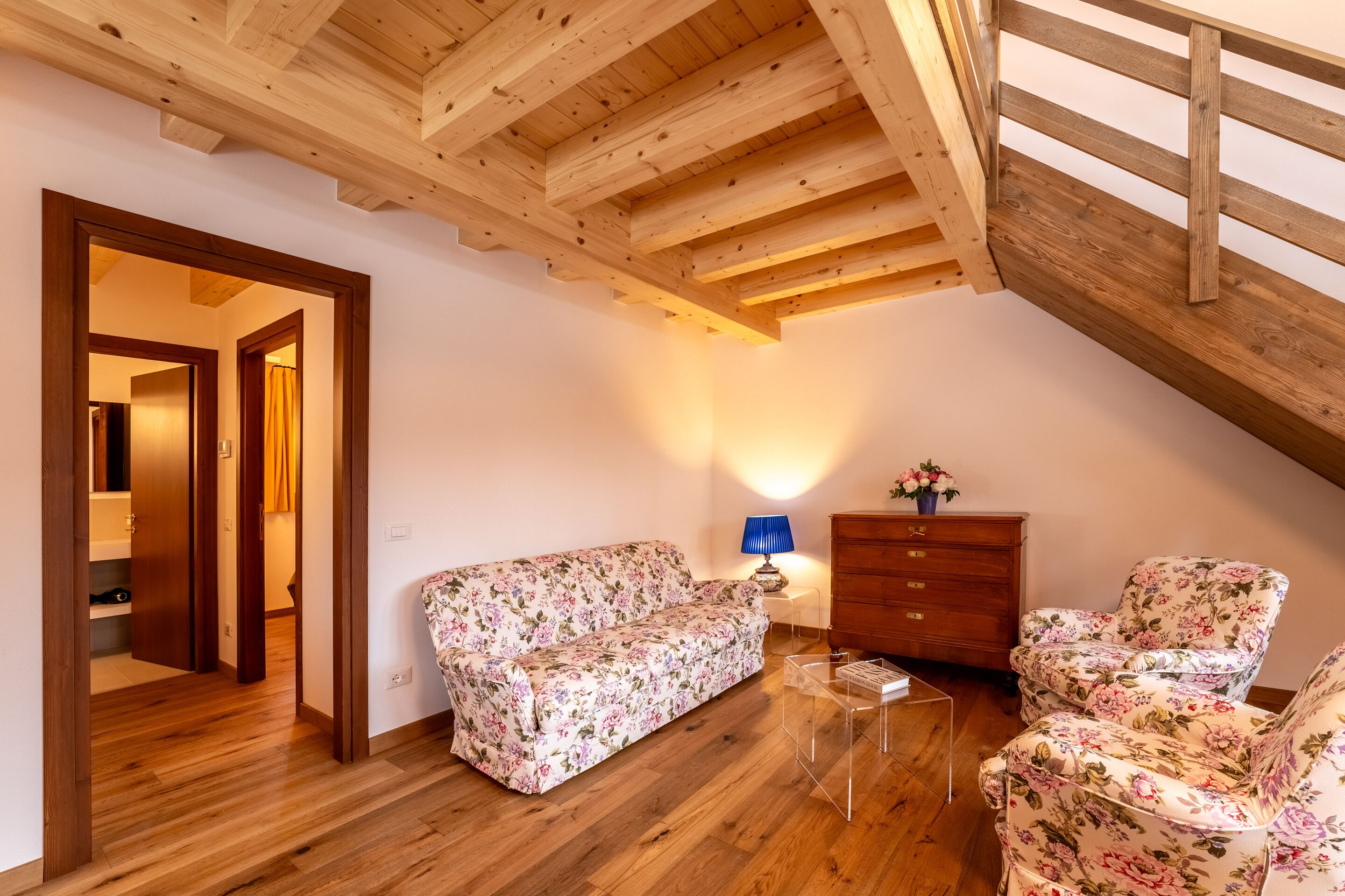Un ambiente accogliente con travi in legno, un divano floreale, una cassettiera di legno e un tavolino da caffè in vetro trasparente.