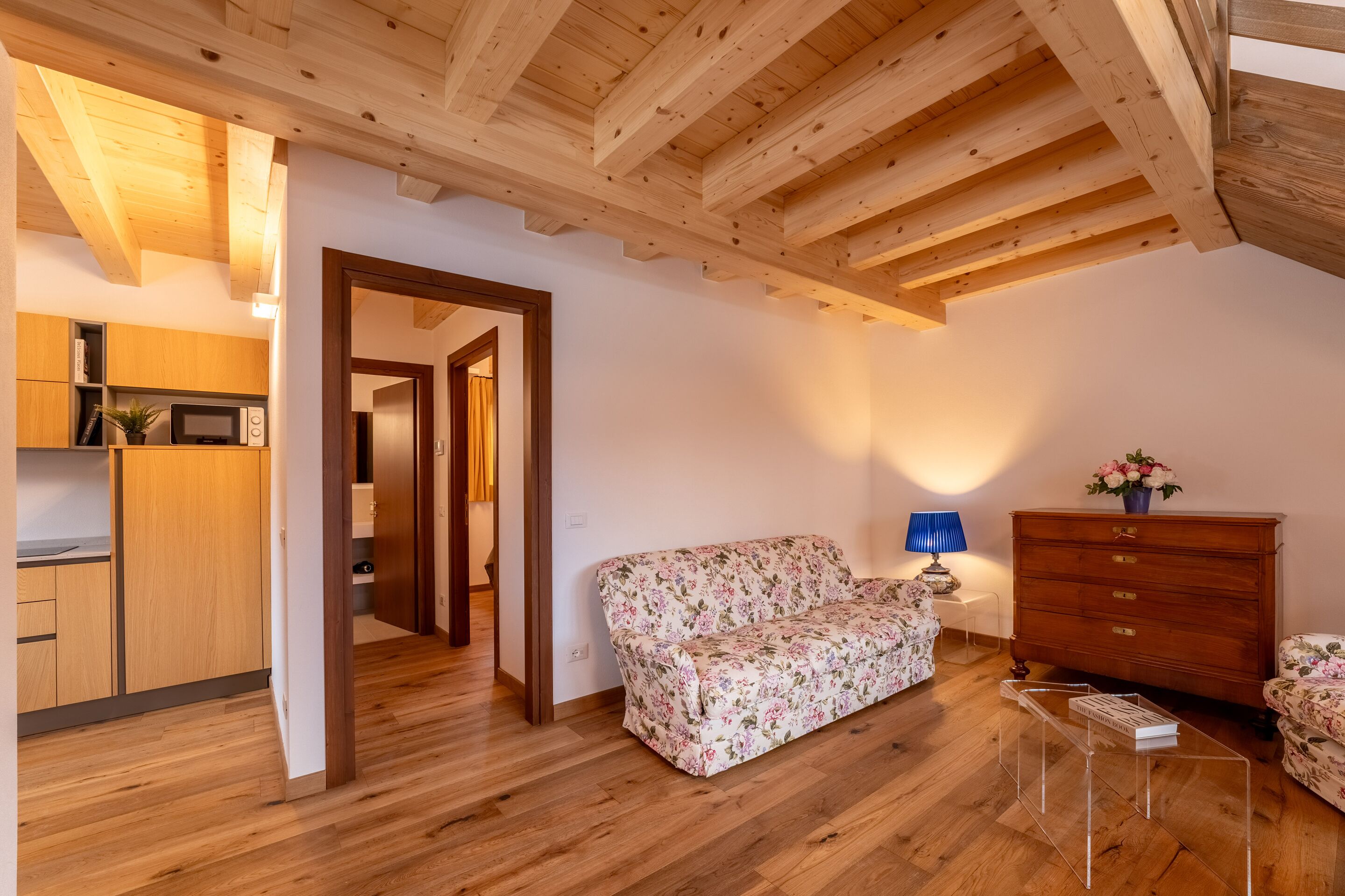 Techo de madera, un sofá floral, cómoda de madera y una mesa de centro de cristal en una habitación con suelo de madera.