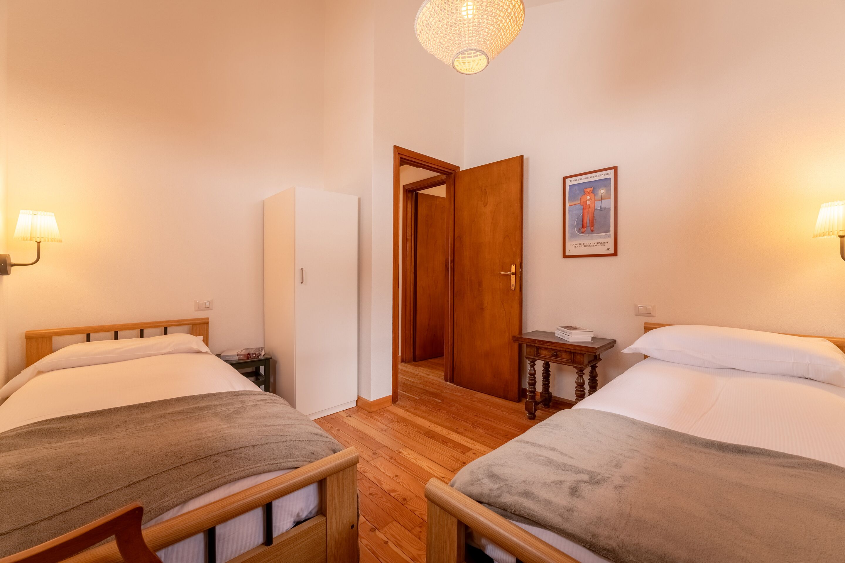 Une pièce avec deux lits, un plancher en bois, une porte donnant sur une autre pièce, une petite table et des lampes fixées au mur.