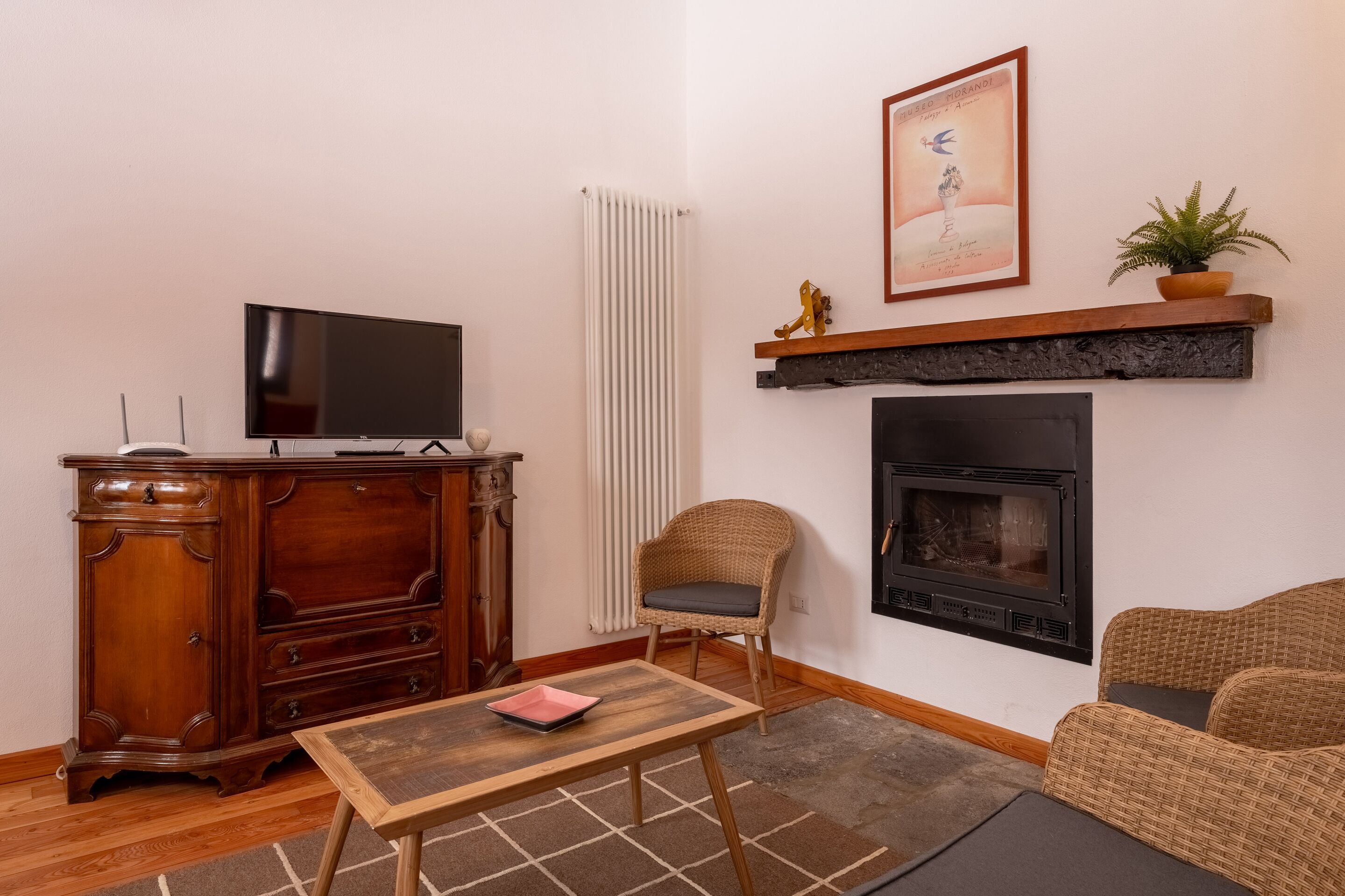 Una sala de estar con televisor, mueble de madera, silla de mimbre, chimenea y un cuadro en la pared.