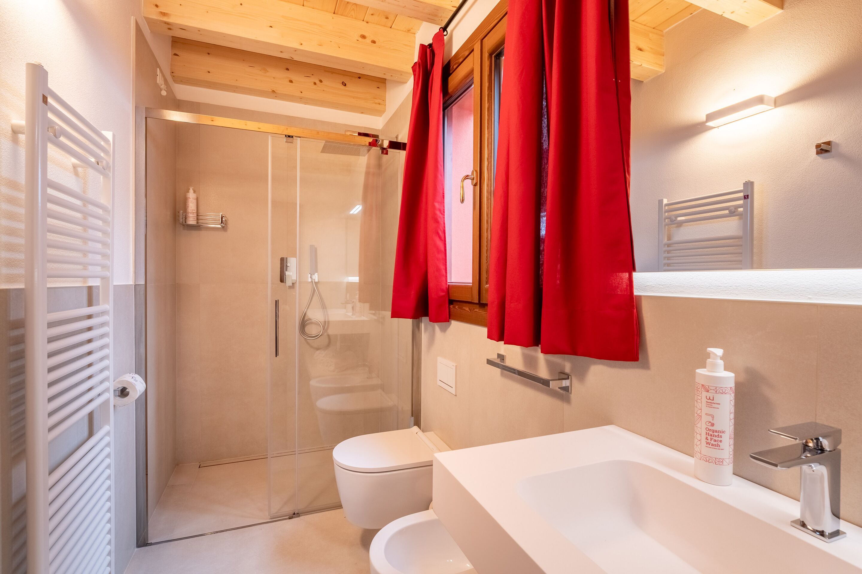 A bathroom with a glass shower enclosure, a white toilet, a sink with a mirror, and a red towel hanging on the door.