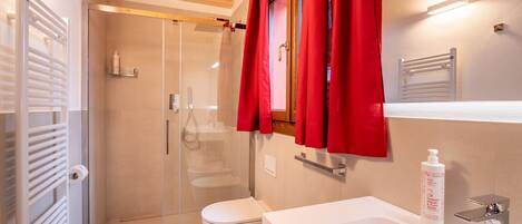 A bathroom with a glass shower enclosure, a white toilet, a sink with a mirror, and a red towel hanging on the door.