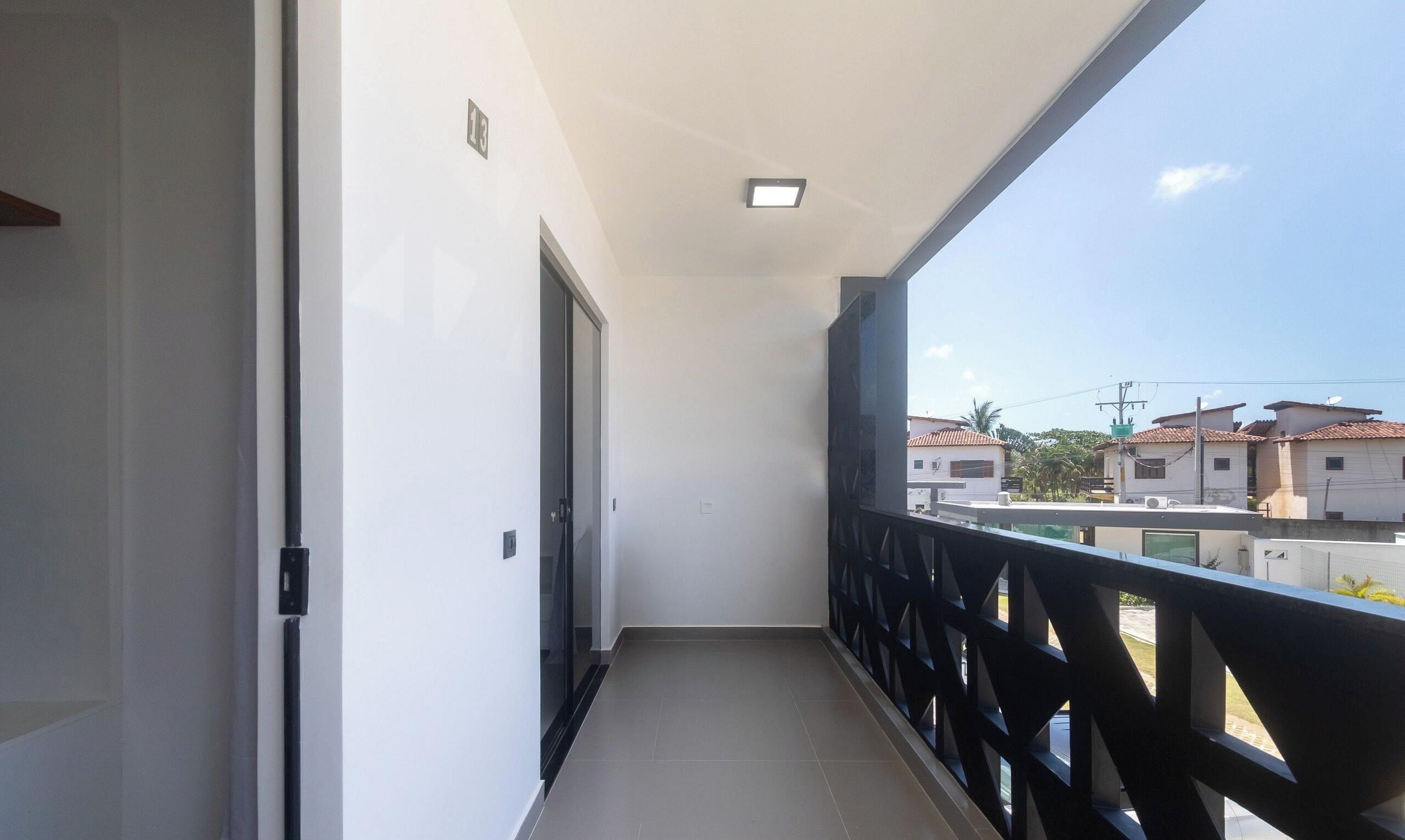 Un balcón estrecho con barandilla negra, una puerta a la izquierda y una vista despejada de edificios al fondo.
