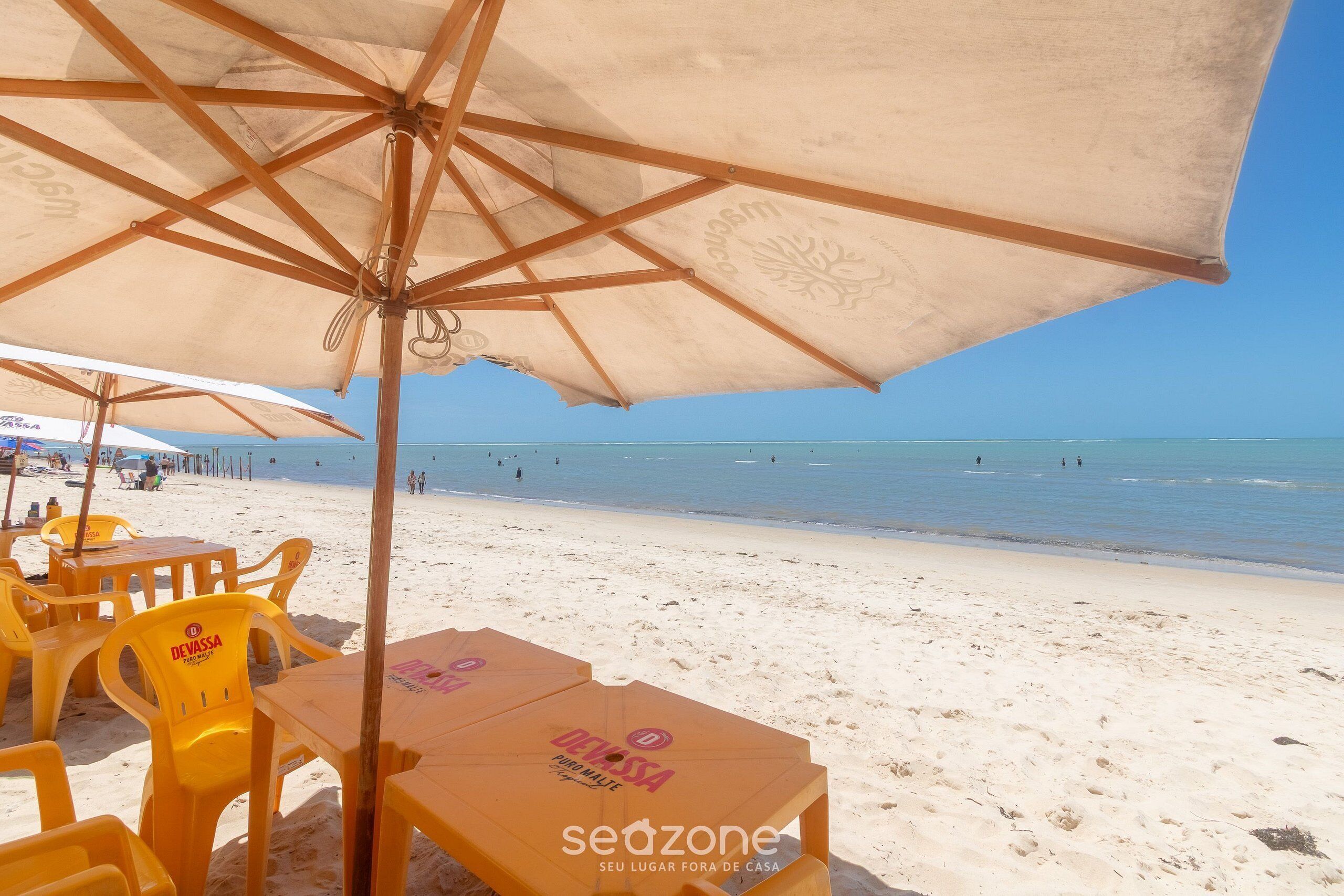 A beachside seating area with yellow chairs and tables under beige umbrellas, with the ocean and beach in the background.