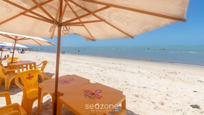 Zona de estar junto a la playa con sillas y mesas amarillas bajo sombrillas color beige, con el océano y la playa al fondo.