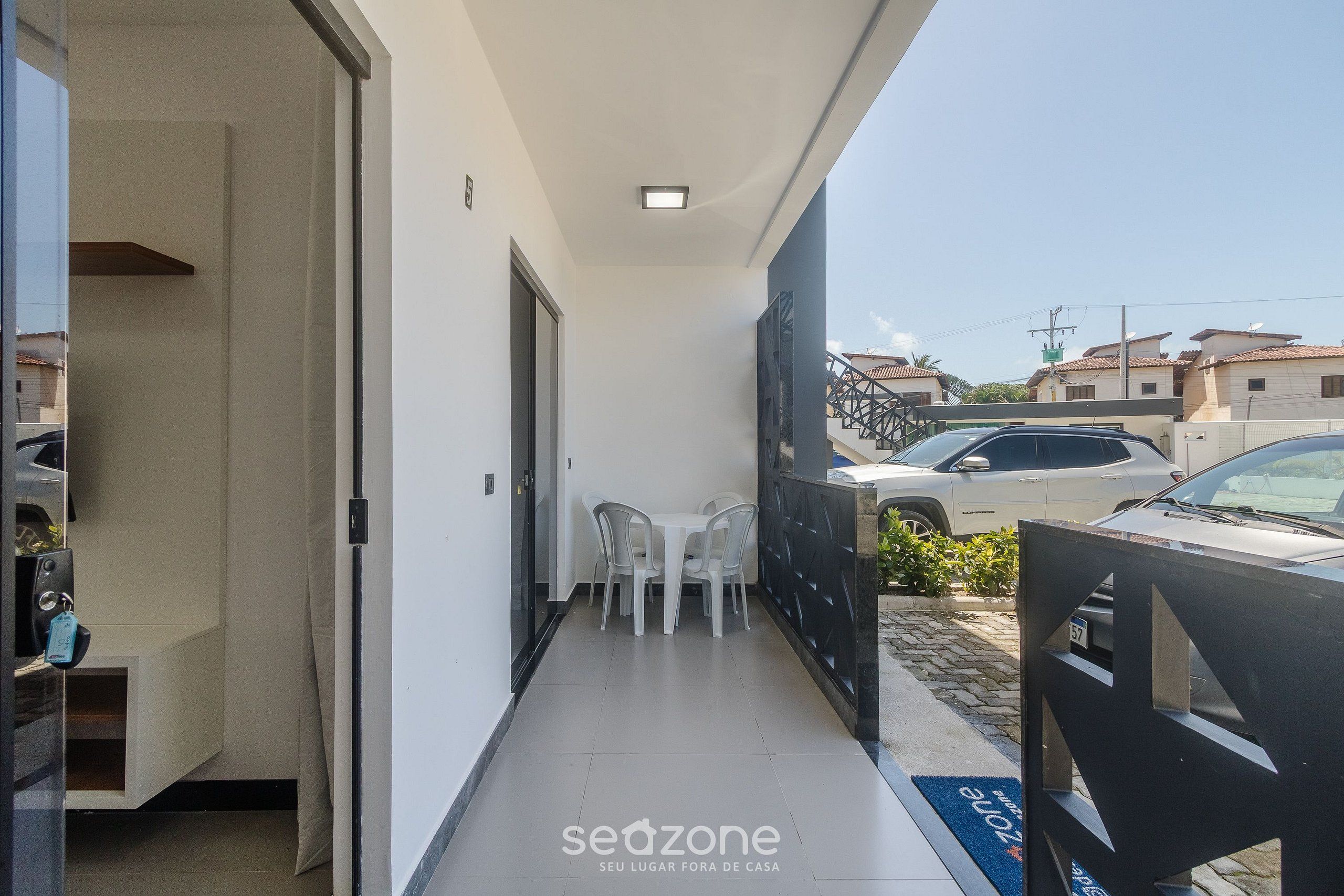 A narrow balcony with a table and chairs, a sliding door, and a view of parked cars and buildings in the background.