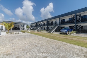 A modern building with a paved courtyard, multiple balconies, and a blue car parked in front. - Apts in a Condo With Barbecue and Pool in the Best Location of Arraial D'ajuda Dvbs (Porto Seguro)