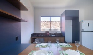 A modern kitchen with a window, a refrigerator, and a dining table set with plates and glasses. - Apts in a Condo With Barbecue and Pool in the Best Location of Arraial D'ajuda Dvbs (Porto Seguro)