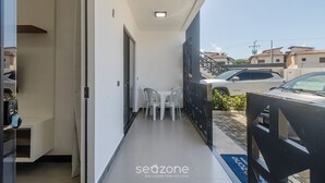 Un balcón estrecho con una mesa y sillas, una puerta corrediza y vistas a coches aparcados y edificios al fondo.