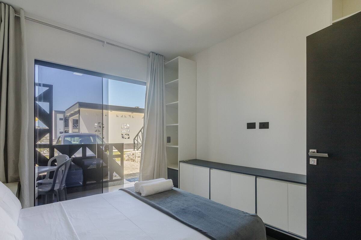 A hotel room with a bed, a small table, a seating area with chairs, a large window, and a view of the outside.