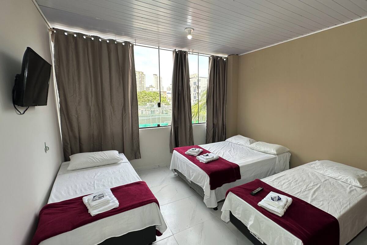 Habitación de hotel con tres camas individuales, un televisor montado en la pared y una ventana con cortinas.