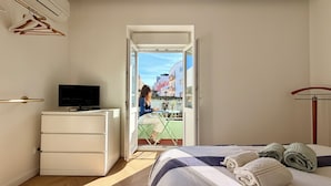 Une chambre à coucher avec un lit, un bureau avec un ordinateur, une télévision et une vue sur la rue par une porte ouverte.