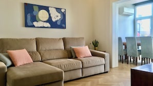 Un salon avec un canapé d’angle beige, une plante en pot et un tableau abstrait bleu accroché au mur.