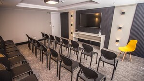 Une salle de conférence avec des rangées de chaises, un grand téléviseur à écran plat et une petite table blanche sur laquelle est posé un vase.