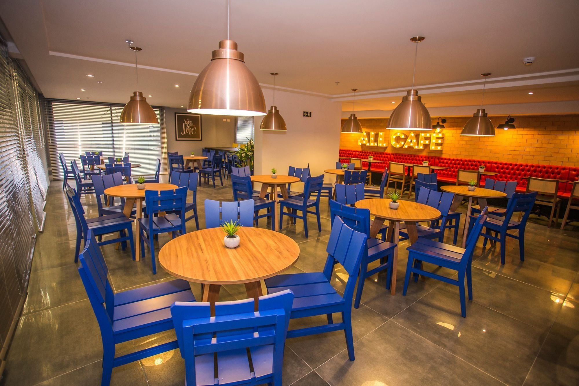 Un café moderno con mesas de madera redondas y sillas azules, con iluminación colgante y una pared de ladrillo rojo de fondo.
