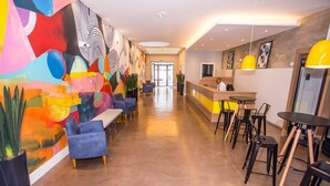 A modern hotel lobby with a long reception desk, blue sofas, and a colorful abstract mural on the left wall. - Studio com Sacada All You Need ALL0305 (Curitiba)