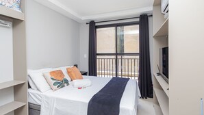 A modern hotel room with a bed, a TV, a balcony with curtains, and a view of the outside. - Studio com Sacada All You Need ALL0305 (Curitiba)