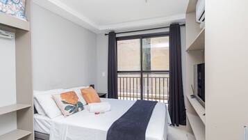 Una habitación de hotel moderna con cama, televisión, balcón con cortinas y vistas al exterior.