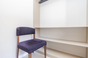 A chair with a dark cushion and wooden legs, positioned against a white wall with a shelf. - Studio com Sacada All You Need ALL0305 (Curitiba)