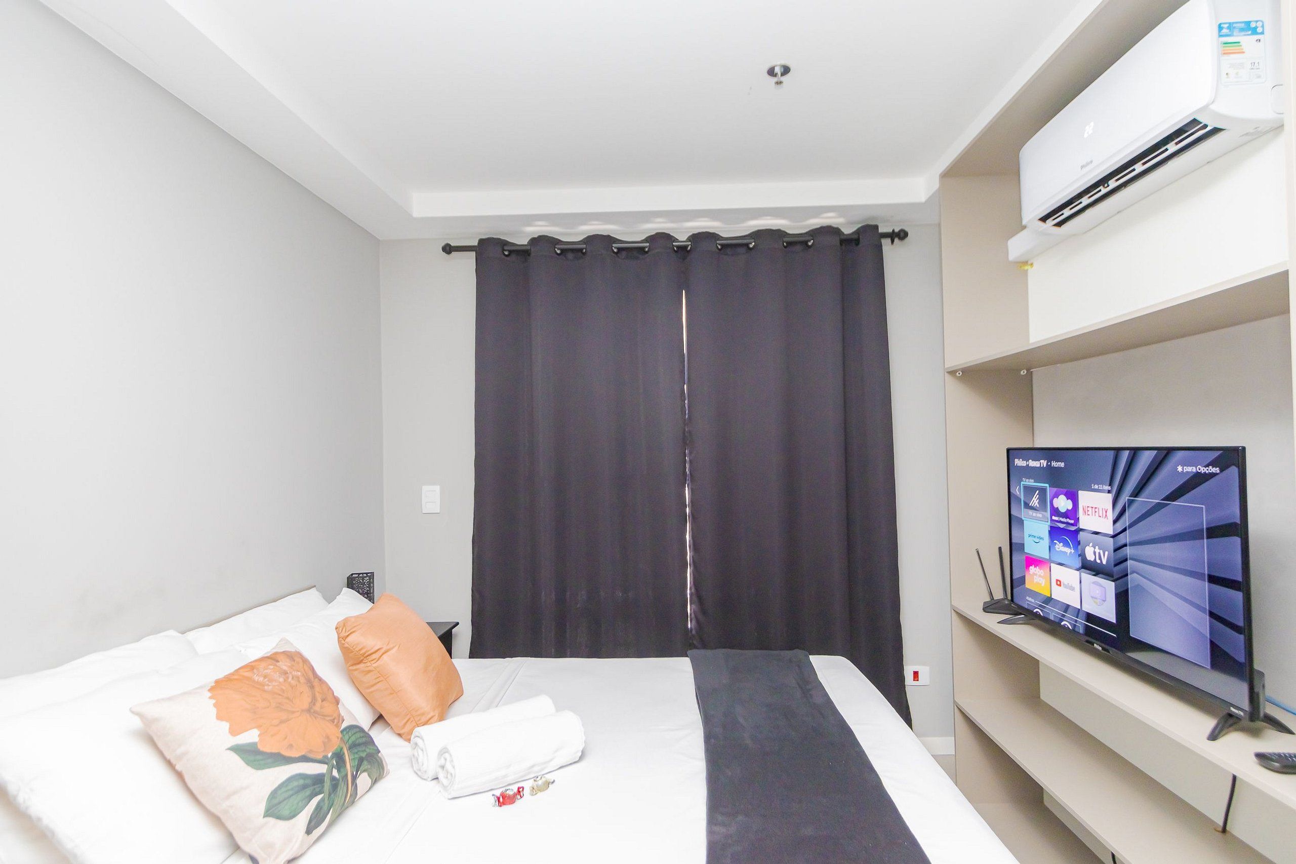 Una habitación de hotel con cama, televisor de pantalla plana, aire acondicionado instalado en la pared y cortina.