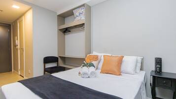 Cama bem arrumada com manta escura, duas almofadas e uma toalha enrolada. Há uma cadeira e uma mesa pequena com um objeto decorativo.