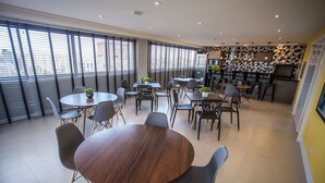 A modern dining area with round tables and chairs, large windows with blinds, and a kitchen area with geometric wall patterns. - Studio com Sacada All You Need ALL0305 (Curitiba)