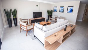 A modern living room with a white sofa, wooden coffee table, and a flat-screen TV. There are two potted plants and a small table with a remote control. - Studio com Sacada All You Need ALL0305 (Curitiba)