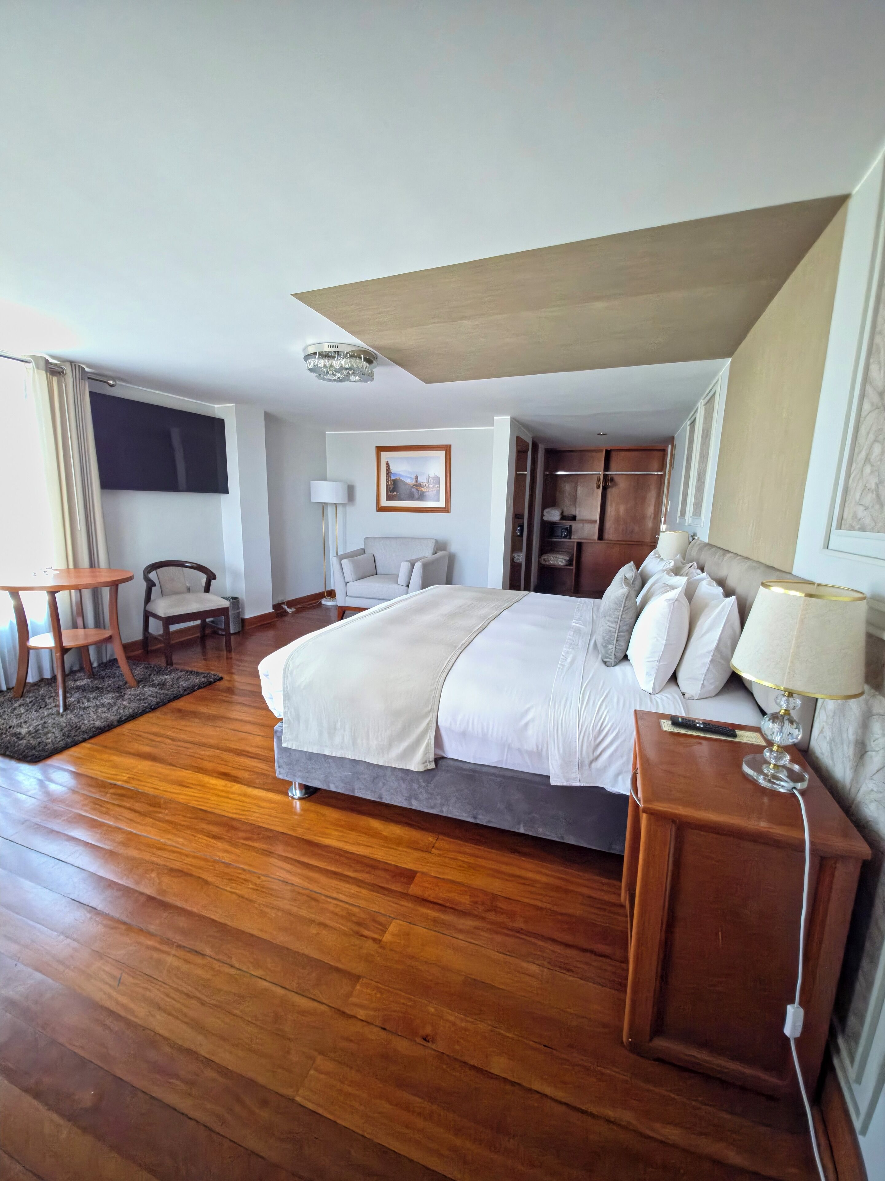 Habitación de hotel con una cama grande, una mesita de noche con lámpara, un escritorio con silla, un televisor y un cuadro en la pared.