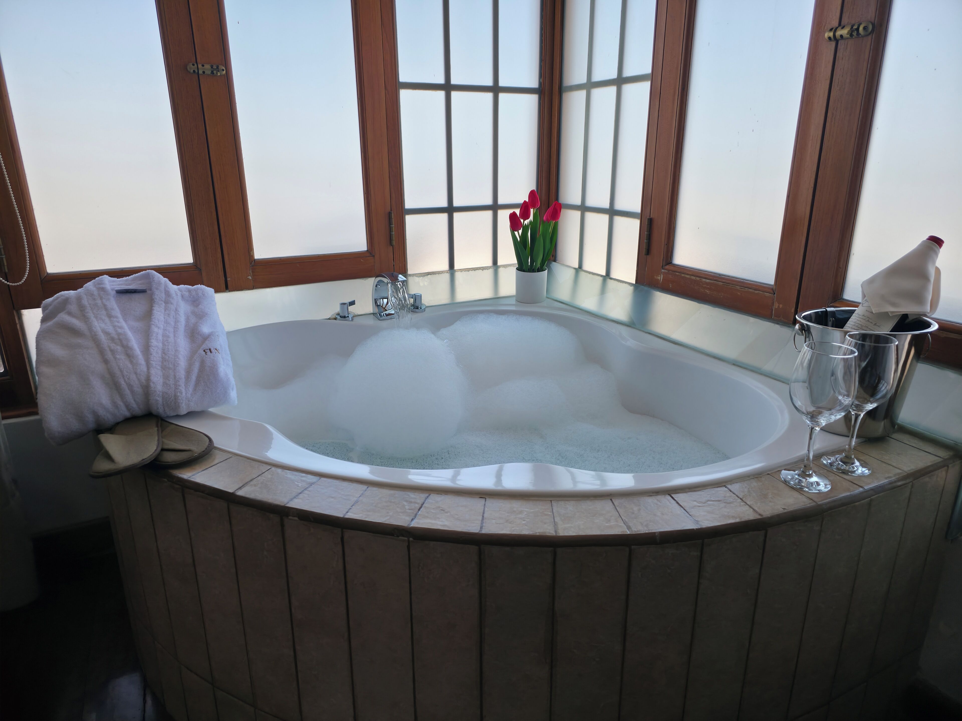 A bathtub with a towel and slippers, a potted plant, and two glasses in a bucket.