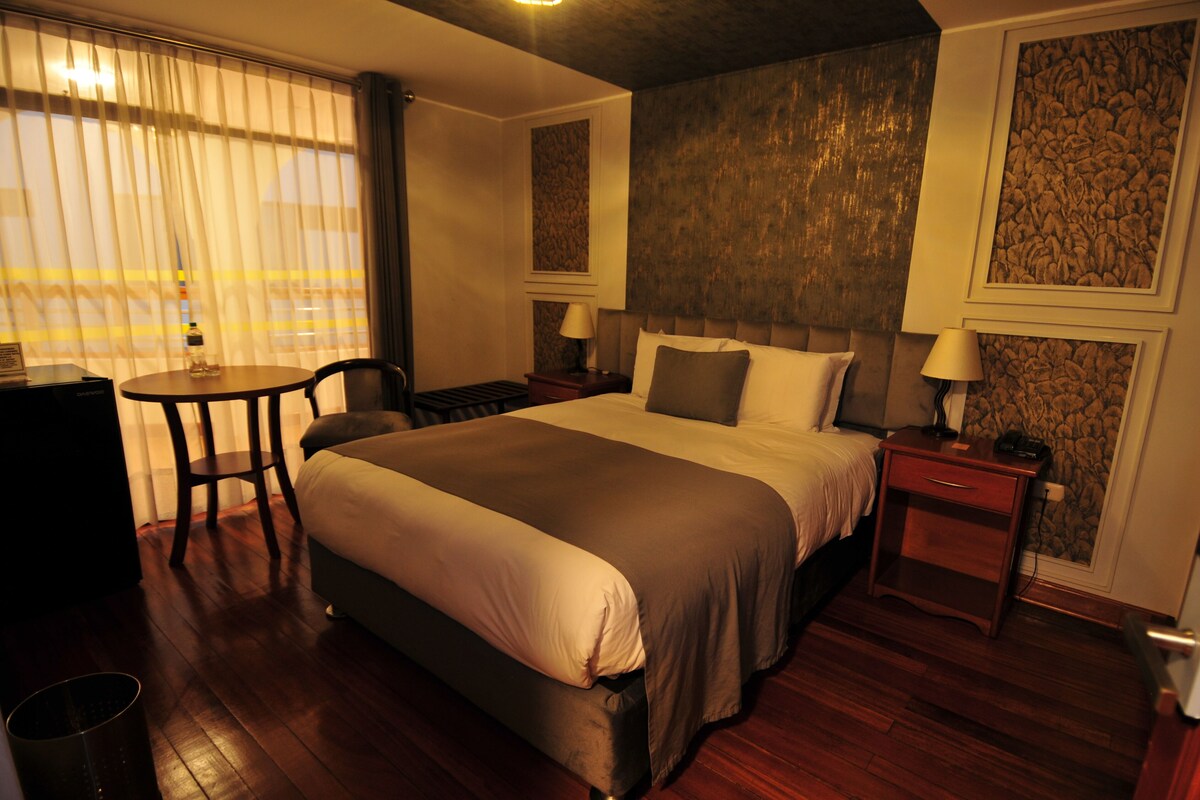 A hotel room with a large bed, a small table, two bedside tables, and a chair.