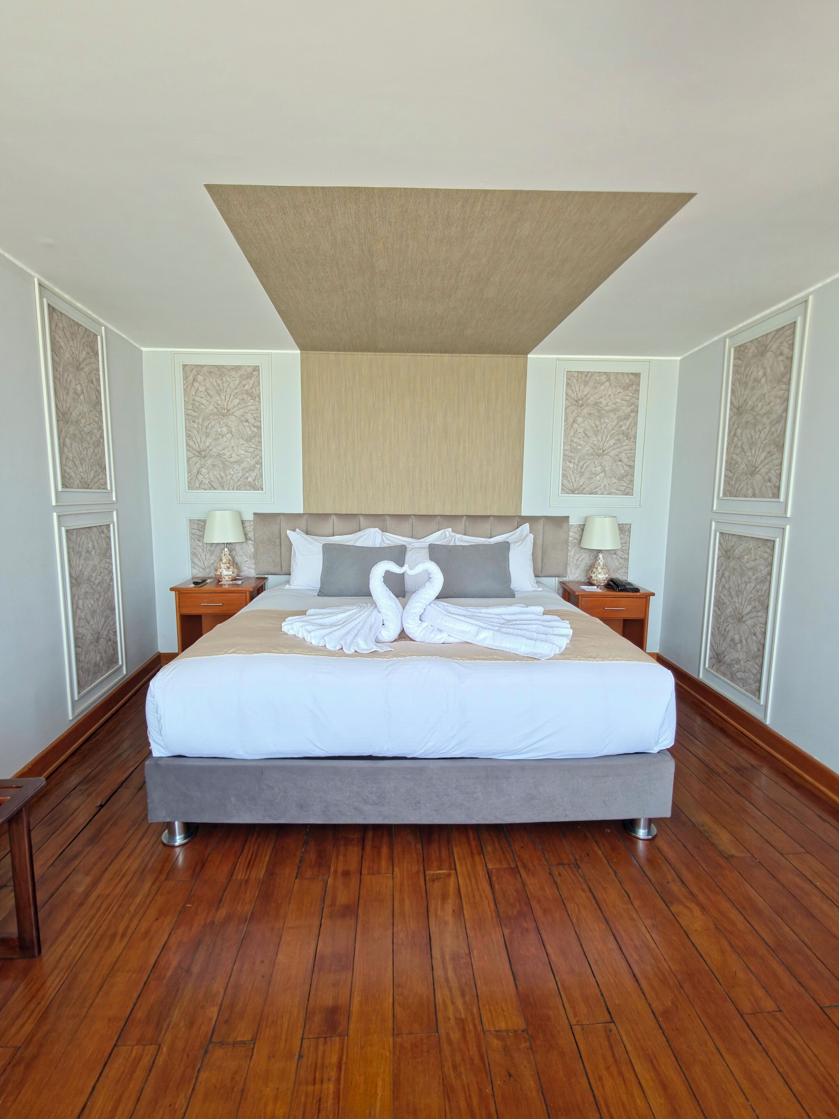 A large bed with a heart-shaped towel arrangement, wooden flooring, and a neutral color scheme.