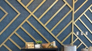 A modern living room with a geometric patterned wall, a wooden cabinet with baskets, a small table with decorative items, a water dispenser, and a grey sofa. - Hotel Fundador By Robles (Arequipa)