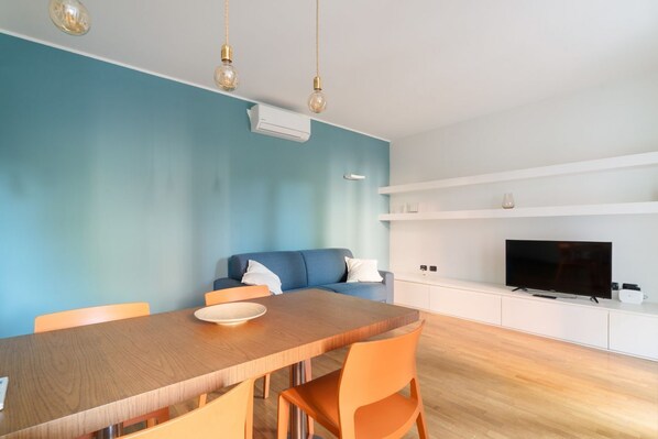 A modern living room with a blue wall, a wooden dining table, orange chairs, a blue sofa, a flat-screen TV, and a white cabinet.