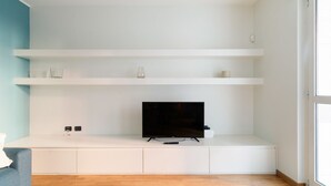 A modern living room with a flat-screen TV on a white console table, wooden flooring, and a large window on the right. - GuestHost - Bright Apartment M1 Precotto Milano (Milan)