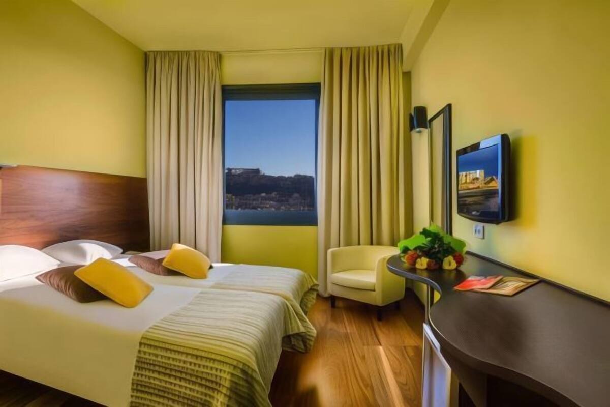 A hotel room with a bed, a desk, a chair, a TV, and a window with curtains.